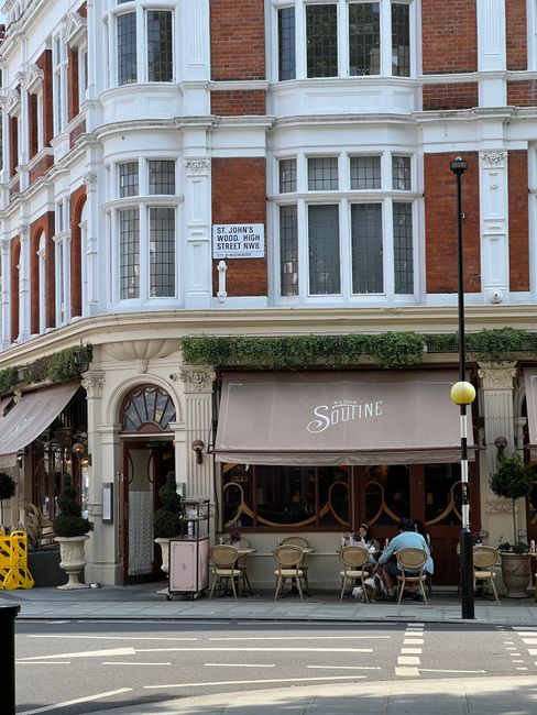 A street view of a multi-story building with large bay windows and detailed architectural features, located on St John's Wood High Street; the ground floor houses a café with a beige awning labeled '